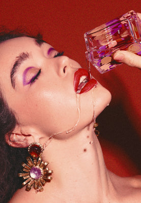 Woman drinking from a pink perfume bottle against a red background