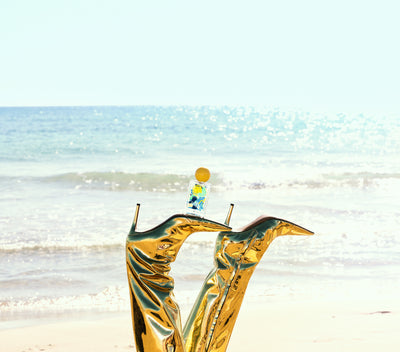 Golden boots with perfume bottle on a beach with a blue sky and ocean in the background