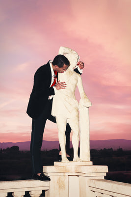 Man in formal attire embracing a statue against a pink sky